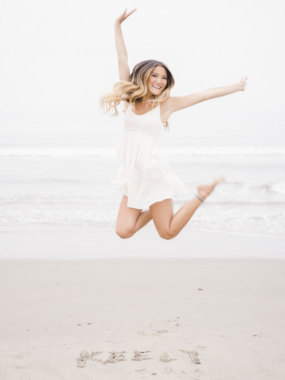 Teenage girl jumping on beach with white dress on and name written in the sand in front of her