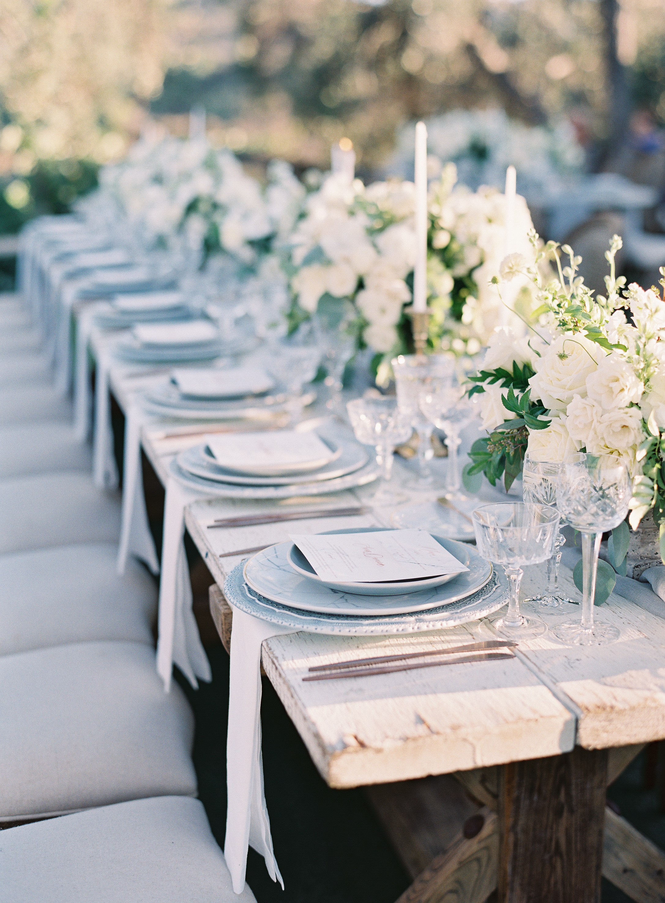 Side view of farm/family style long table setting.  White rose and mixed white flower center pieces.  Rustic/modern