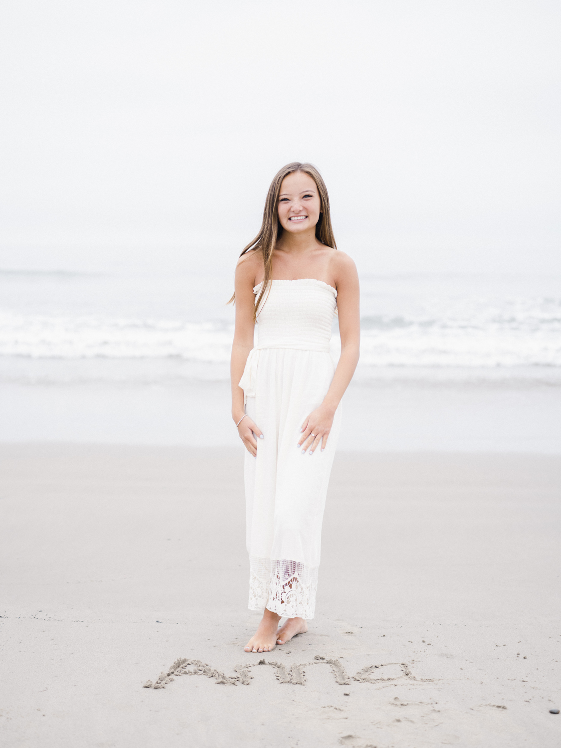 Girl in white standing on the beach with her name written in the sand at her feet