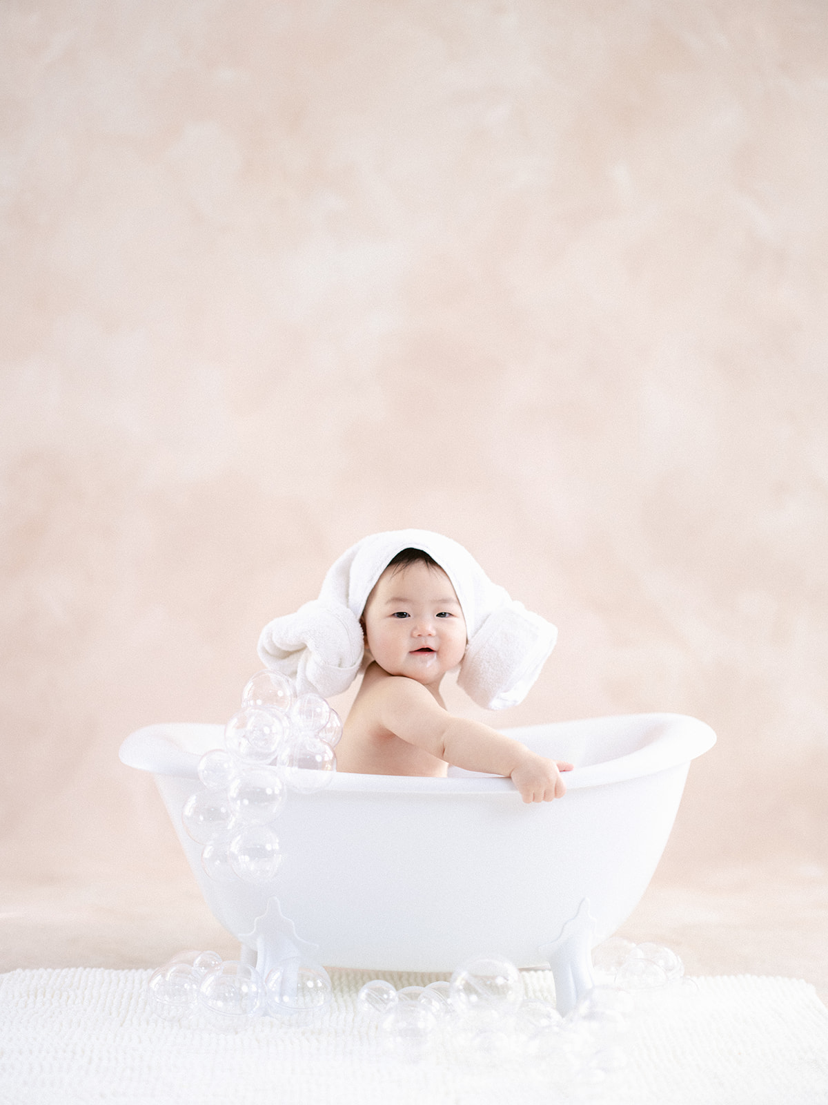 spa day with cute asian baby sitting in a baby-sized clawfoot tub with head wrapped in a towel